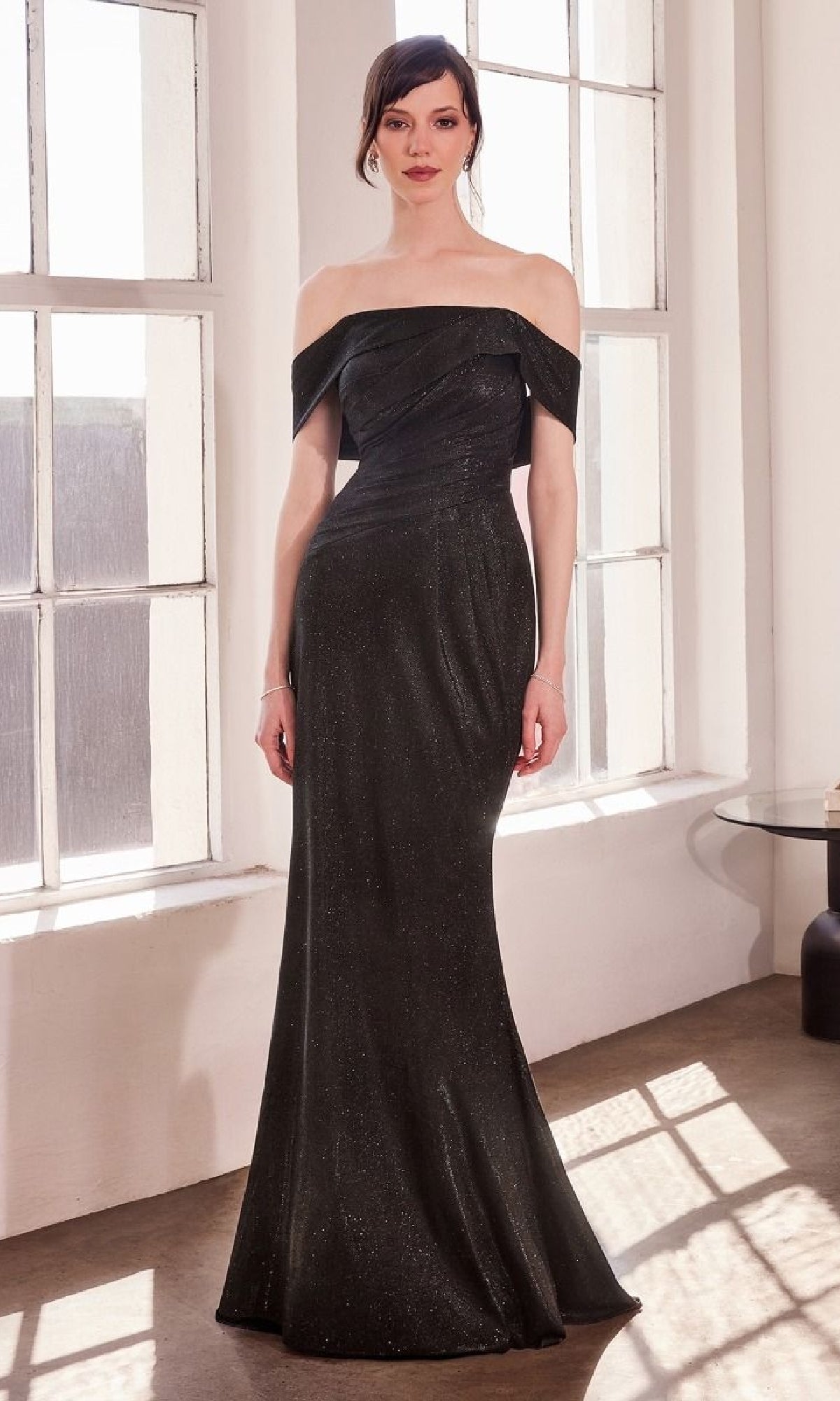 A woman stands in a bright room with large windows and a round glass table, wearing the Amoris T603 Off-Shoulder Long Glitter Prom Dress with a mermaid skirt.